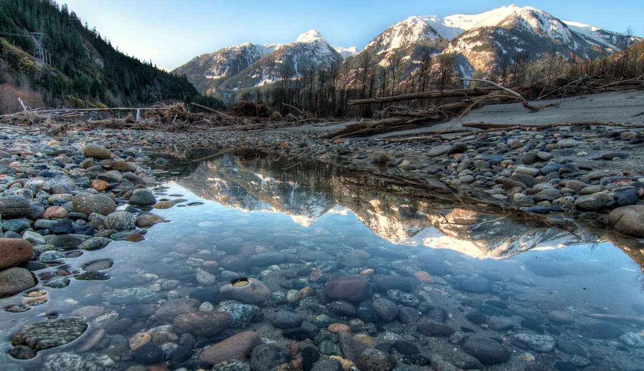 Tranquil winter scene with snow-capped mountains reflected in a serene stream, framed by stones and pebbles.